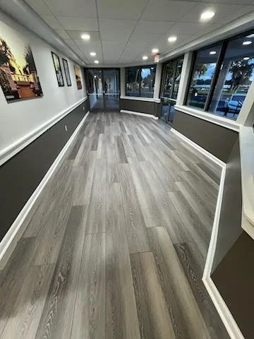 Empty hallway with large windows, wood flooring, white walls with black and white pictures, and ceiling lights.