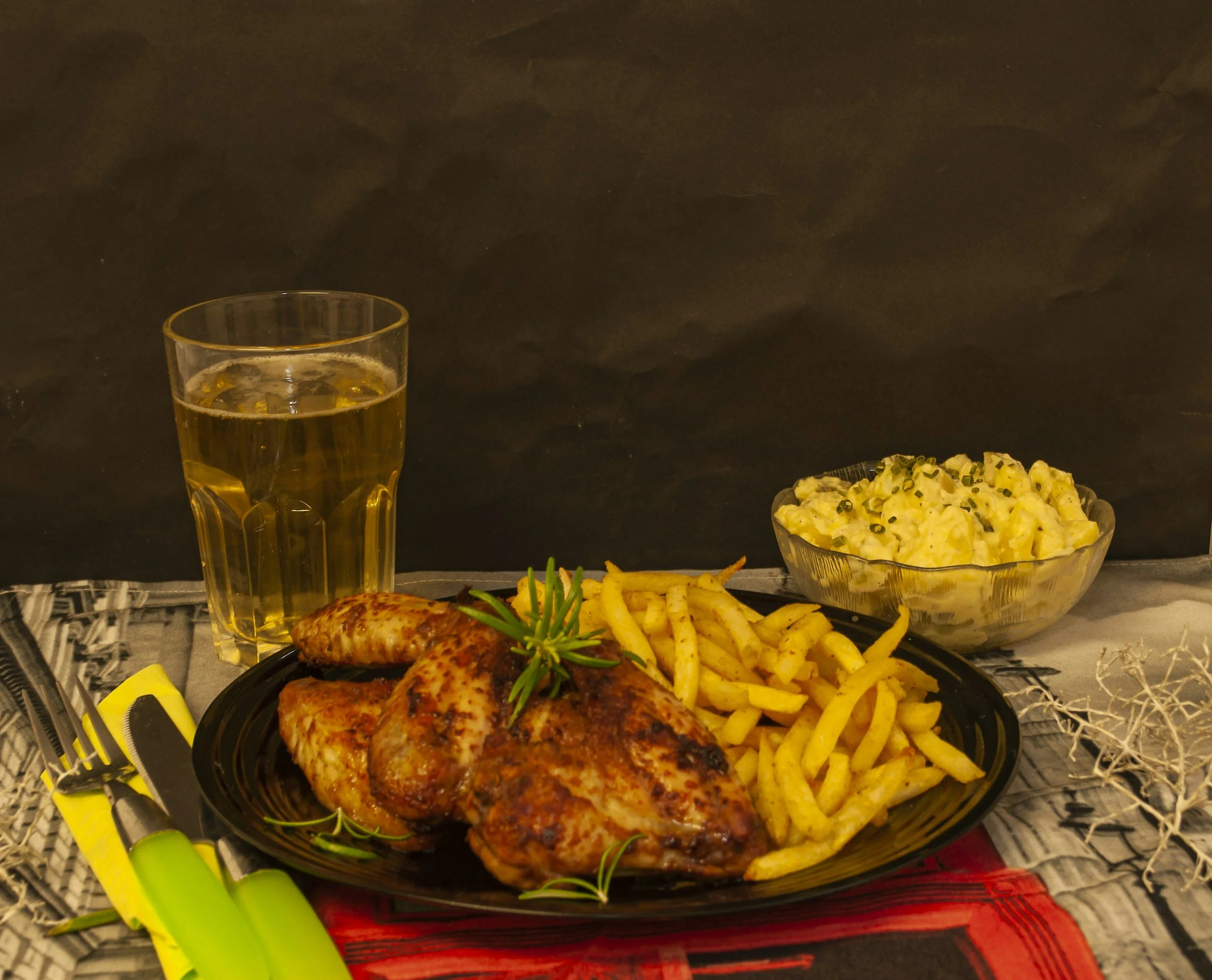A black plate of cooked chicken with herbs, a side of French fries, and a sprig of rosemary. In the background, a bowl of macaroni salad with chopped chives, a glass of iced tea, and additional food items are visible on a table setting with colorful 