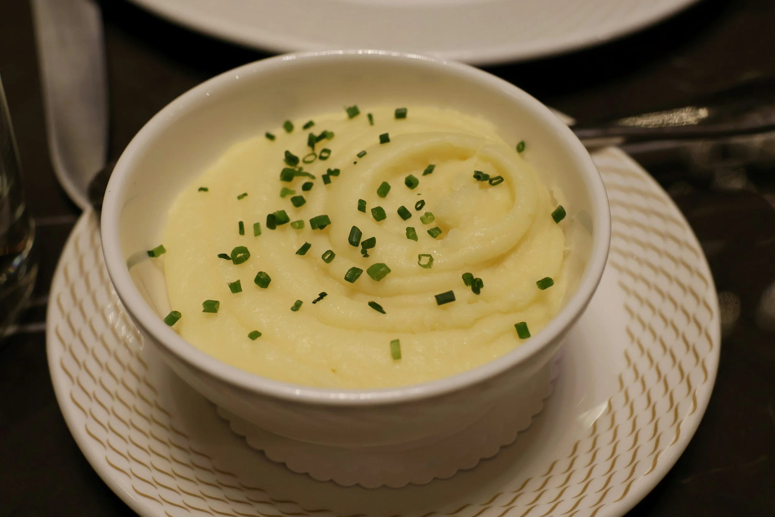 A cup of mashed potatoes topped with chopped chives on a small white dish with a matching cup.