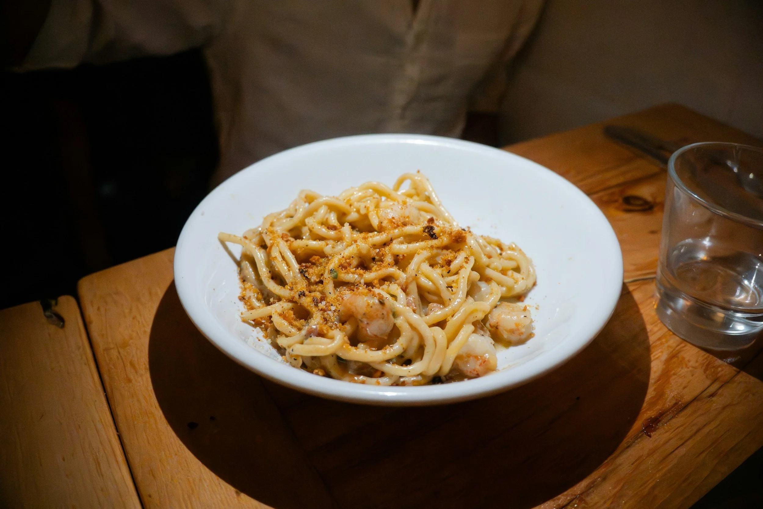 A white bowl of creamy pasta with seasoning on top, placed on a wooden table with a glass of water nearby.