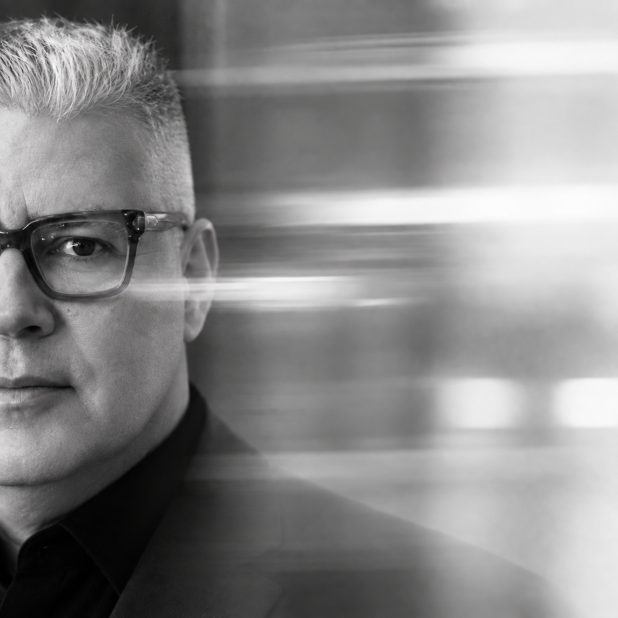 A black and white photo of a man with glasses, looking directly at the camera, with streaks of light or motion blur extending to the right.