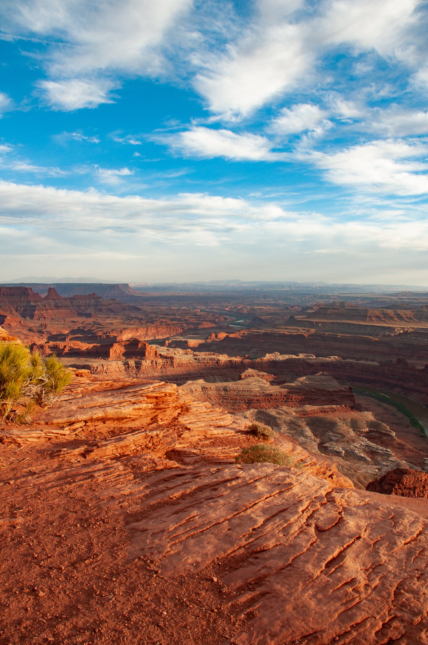 View of the American West with layered red rock formations and a wide river running through the canyon, under a partly cloudy sky.
