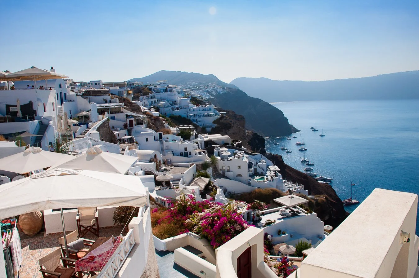White buildings and terraces on a hillside overlooking the sea with boats, pink flowers, and a clear blue sky.