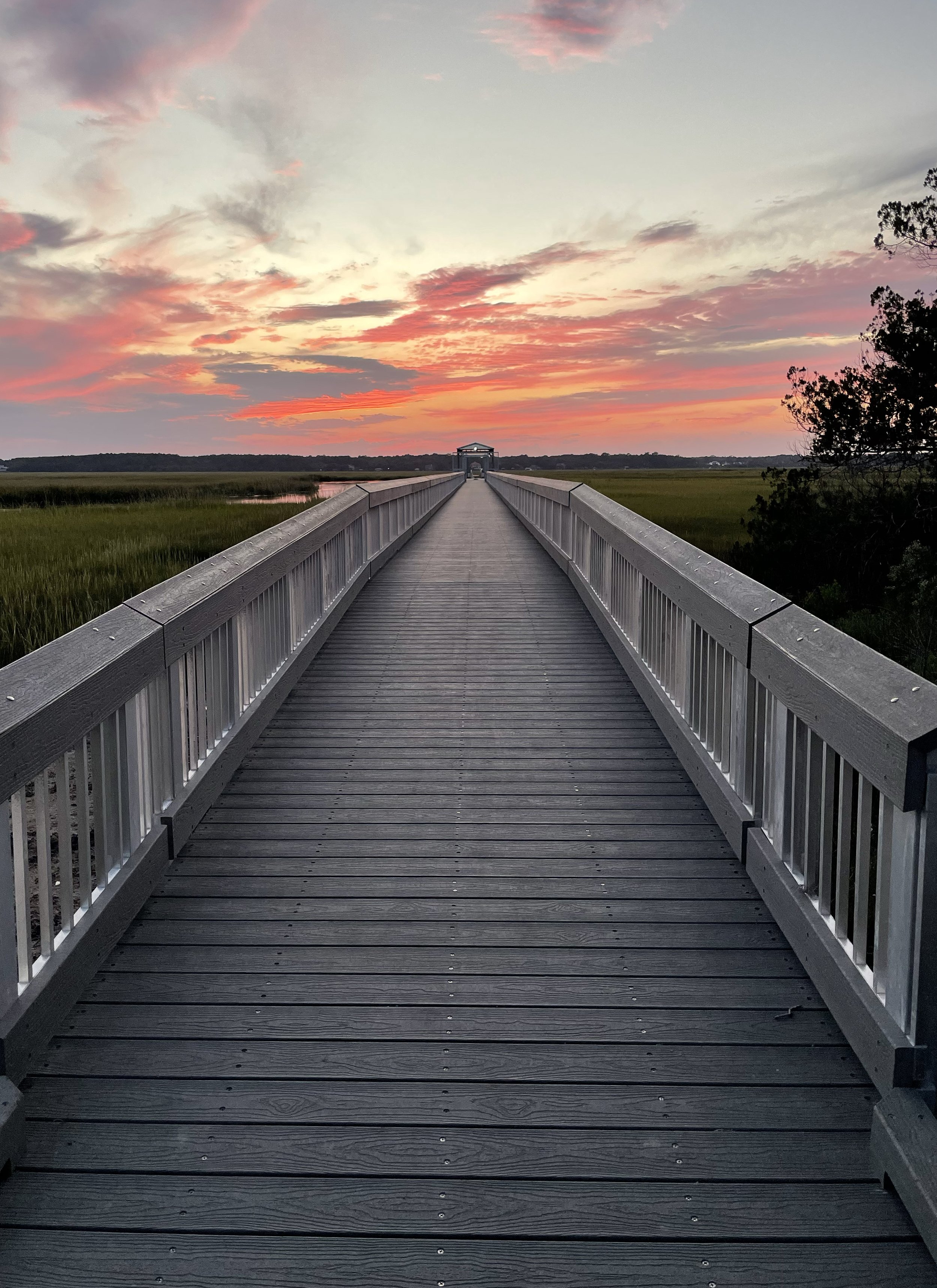 A long wooden pier extends over marshland toward a small covered shelter at sunset, with pink and orange clouds in the sky.