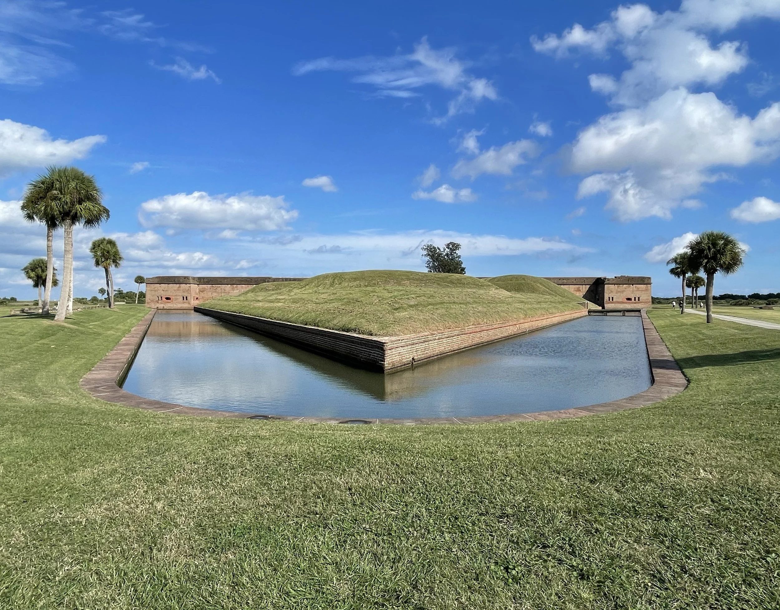 A landscape with a moat surrounding an island with grassy mounds and historic brick structures. Tall palm trees and a bright blue sky with scattered clouds are visible.