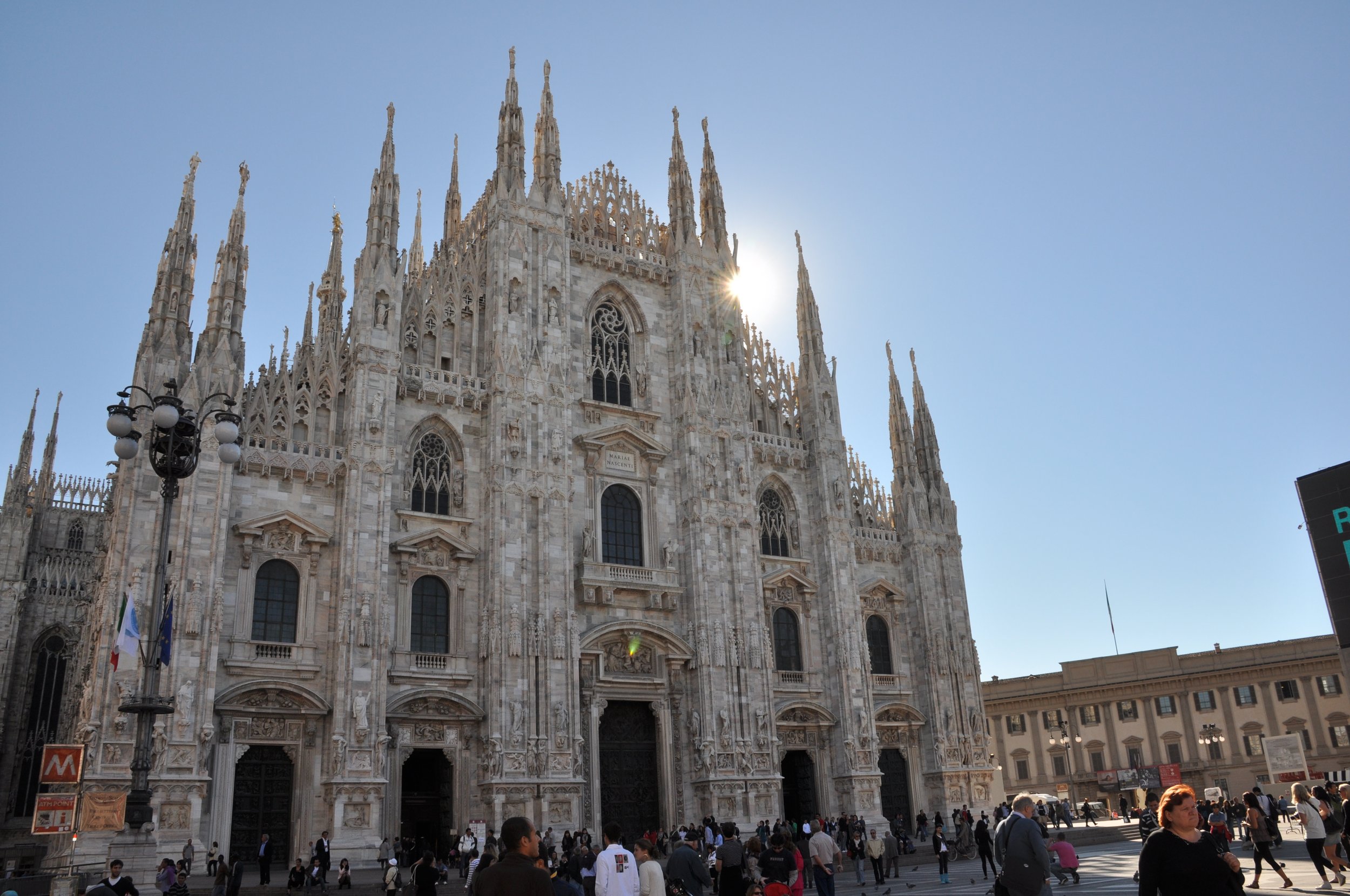 Exterior view of Milan Cathedral (Duomo di Milano), a cathedral with ornate gothic architecture and numerous spires, under a clear blue sky with sunlight peeking through the spires. People are gathered at the base, some walking or standing.