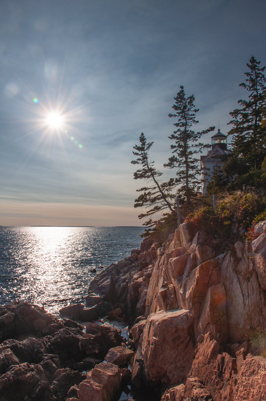 Sun shining over a rocky coastline with a lighthouse and pine trees, reflecting on the water.