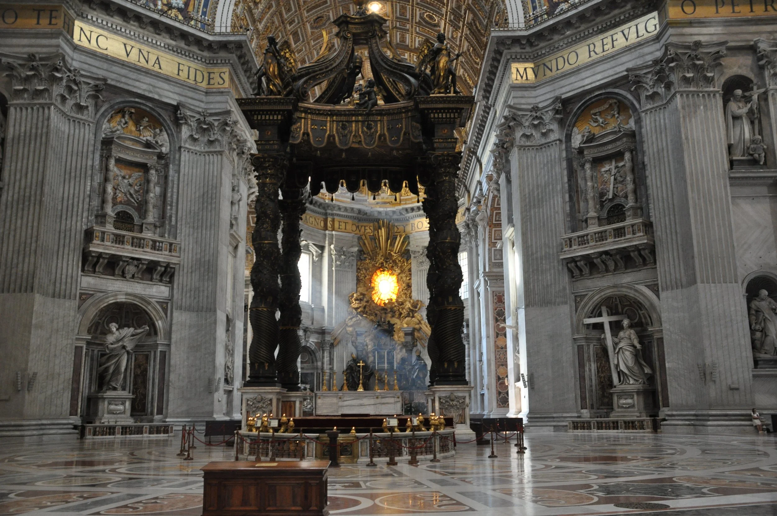 Interior of a grand, ornate church or cathedral with detailed sculptures, columns, and gold accents, featuring a central altar with a gold tabernacle and a large icon of a sun-like structure, lit by natural light.