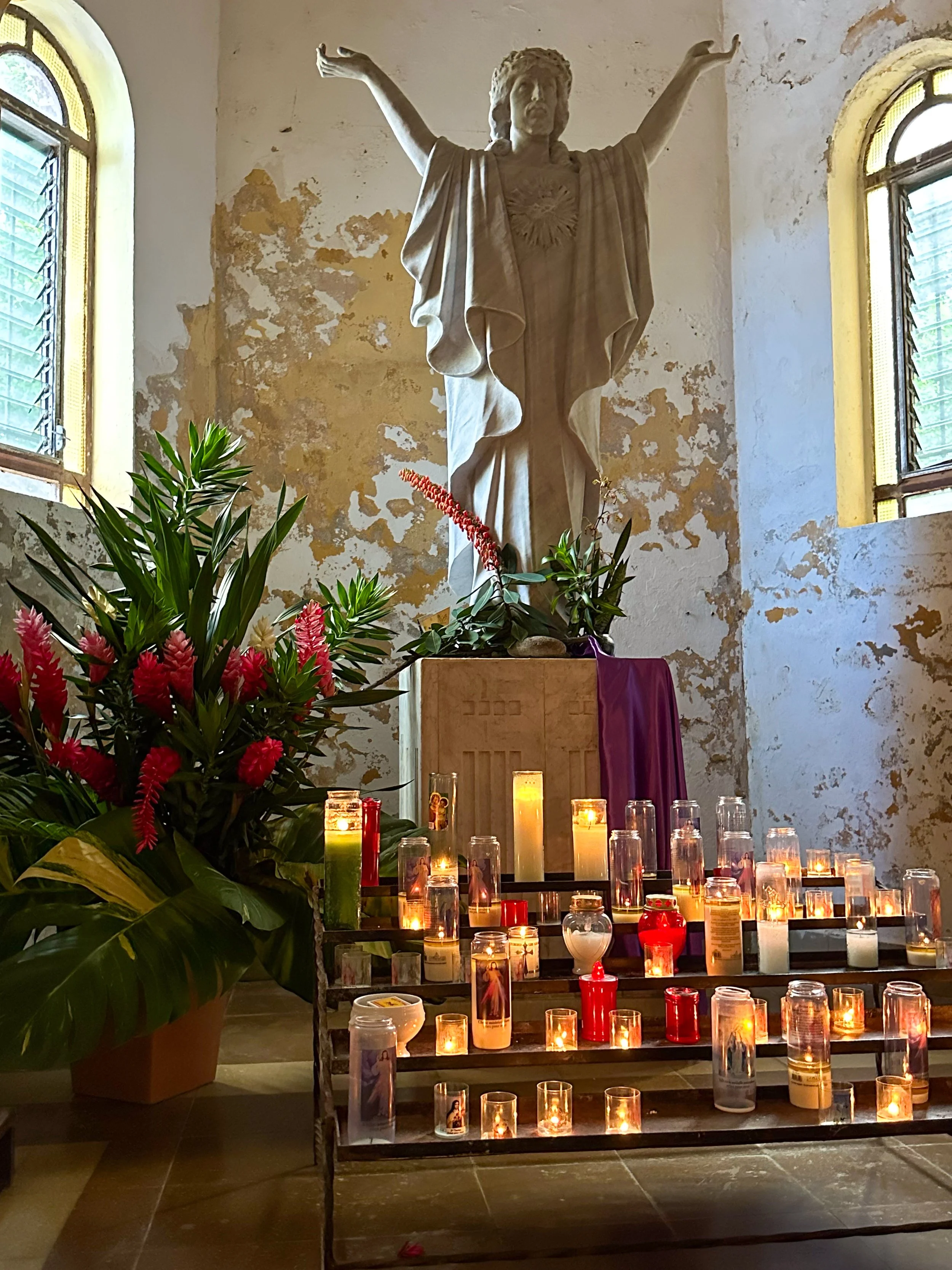 A religious altar with numerous lit candles, a large plant, and a statue of Jesus Christ with arms outstretched, inside a church with peeling paint walls and stained glass windows.