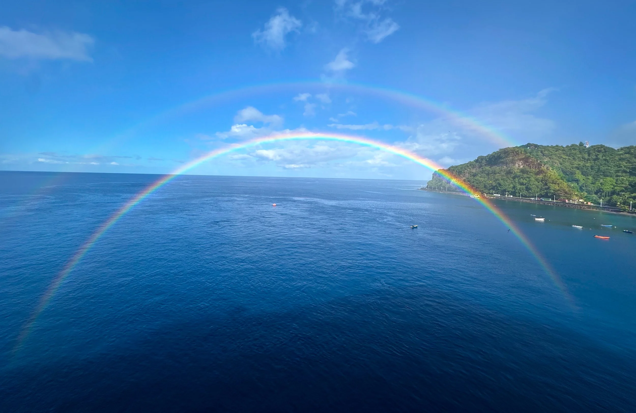 Two rainbows arch over a large body of water with a shoreline covered in green trees and a hill in the background.