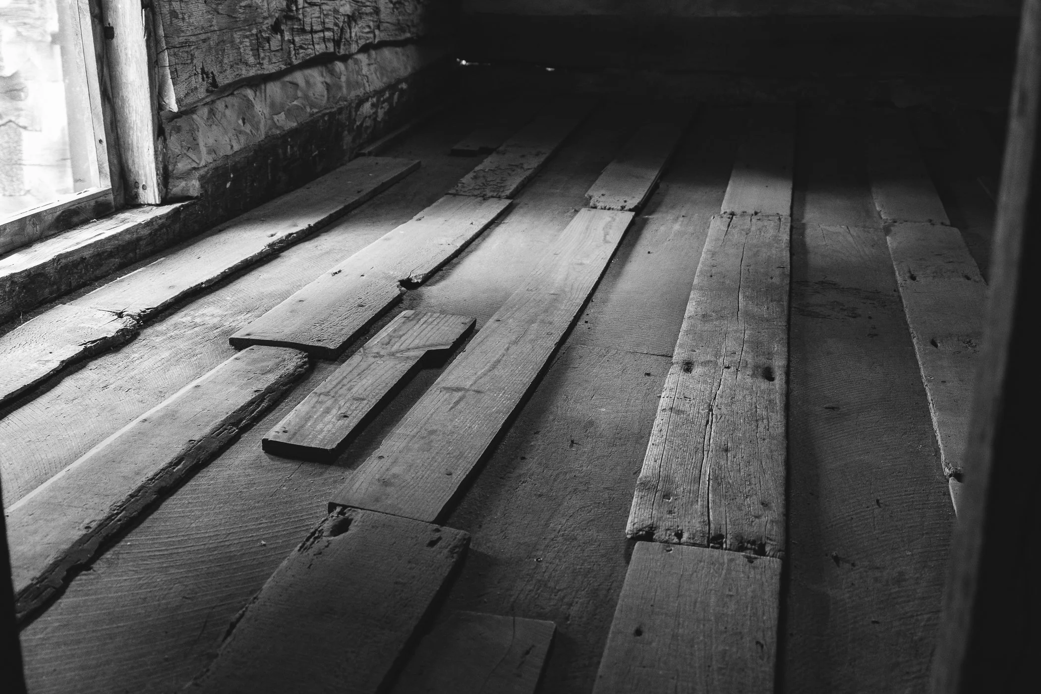 Black and white photo of rough wooden planks and beams inside a rustic building, with a small window letting in natural light.