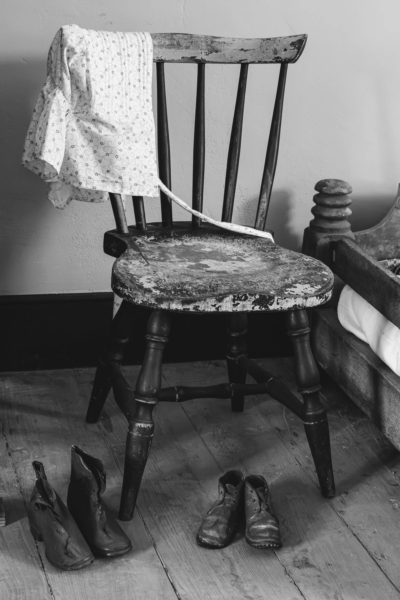 Old wooden chair with a drumstick paint or stain on it, draped with a patterned bonnet, and several pairs of worn work shoes underneath. The setting appears to be indoors with a wooden floor.