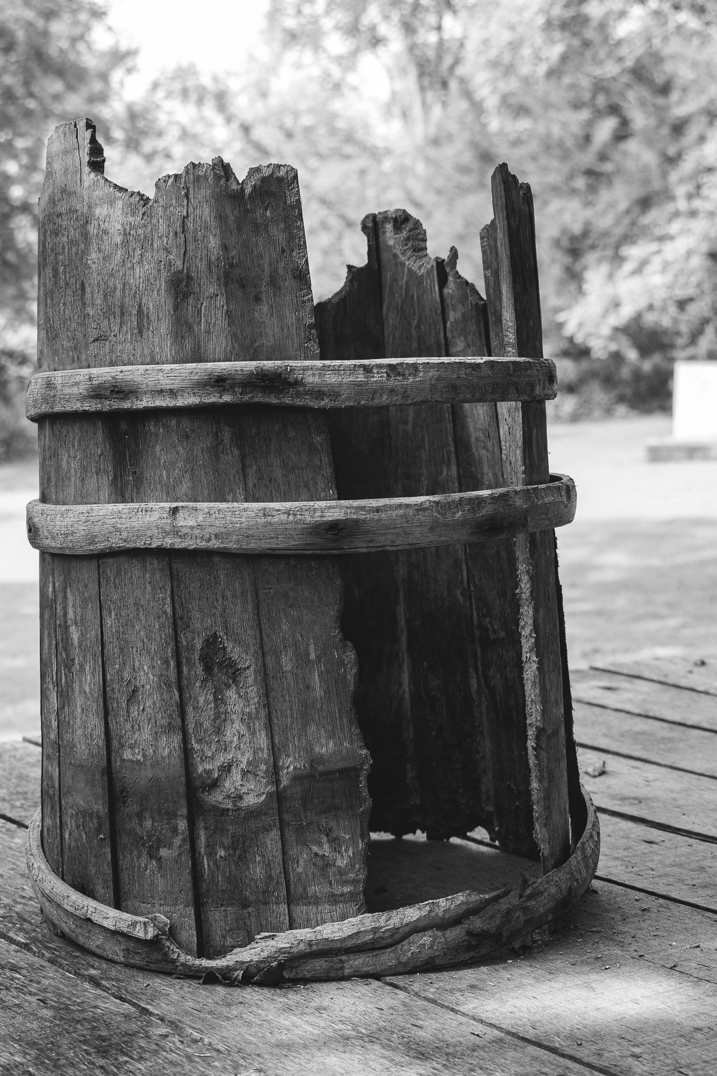 Black and white photo of a weathered, old wooden butter churn with a piece of wood on top, placed on a wooden deck with trees in the background.