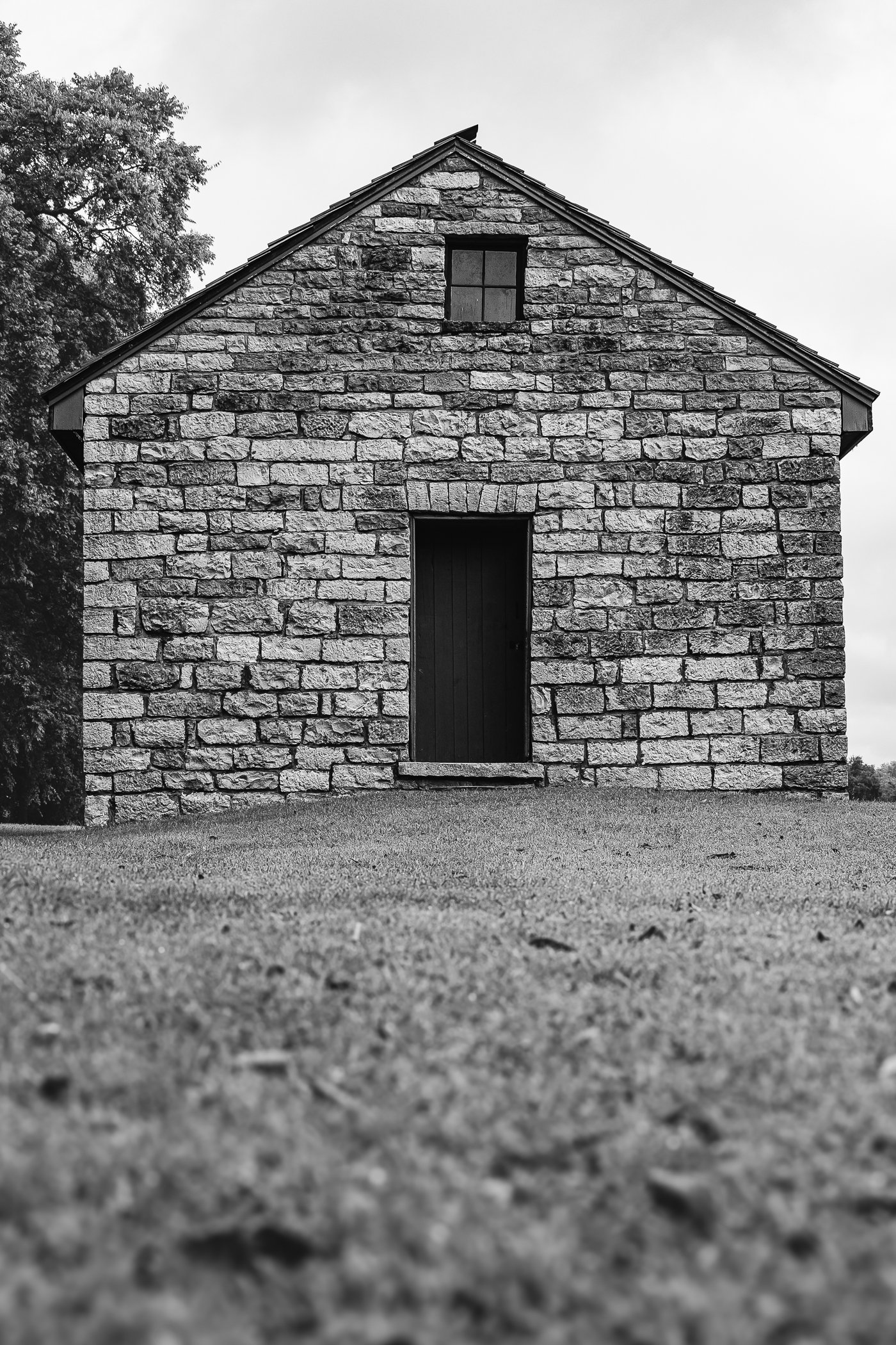 A black and white photo of an old stone building with a pitched roof, a small window near the top, and a boarded-up door at ground level; trees are visible in the background.
