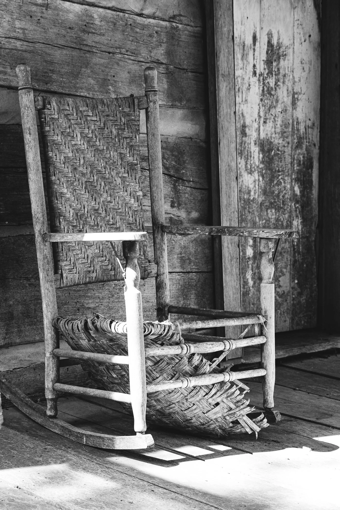 An old wooden rocking chair with woven seat and backrest sitting on a wooden porch, casting a shadow on the floor in black and white.