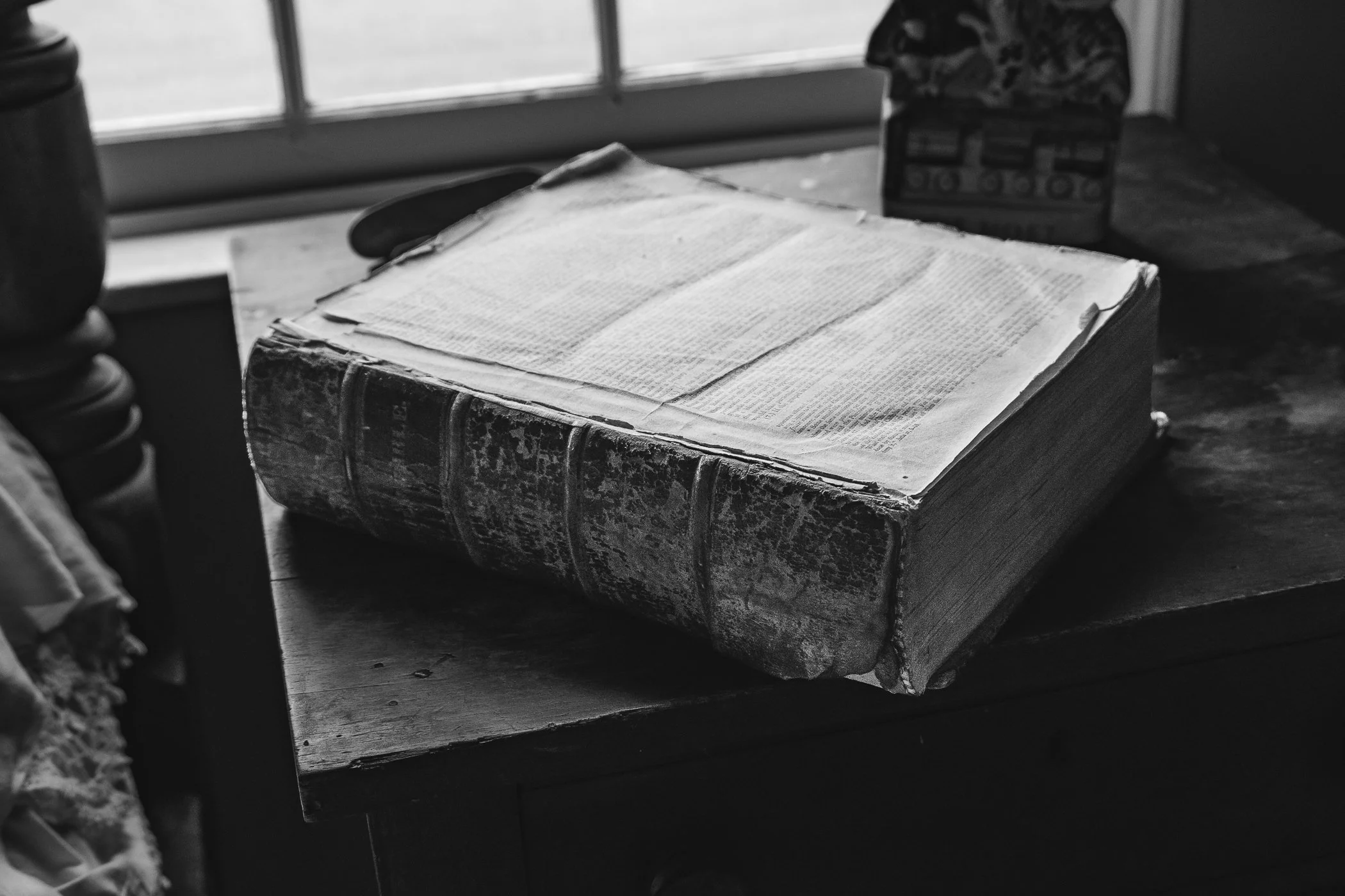 An old, large Bible with a worn leather cover resting on a wooden table near a window with natural light. In the background, there is a decorative box and a bottle, possibly a soda or water bottle. The room has a vintage or rustic feel.