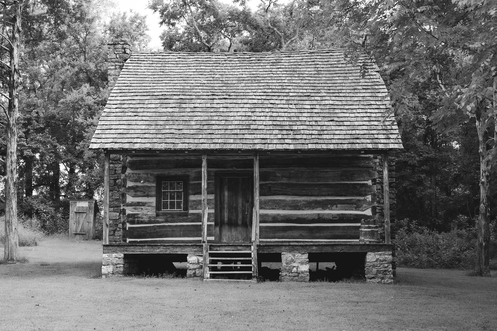 Black and white photograph of a rustic, old wooden cabin with a steep shingled roof, surrounded by trees and grass.