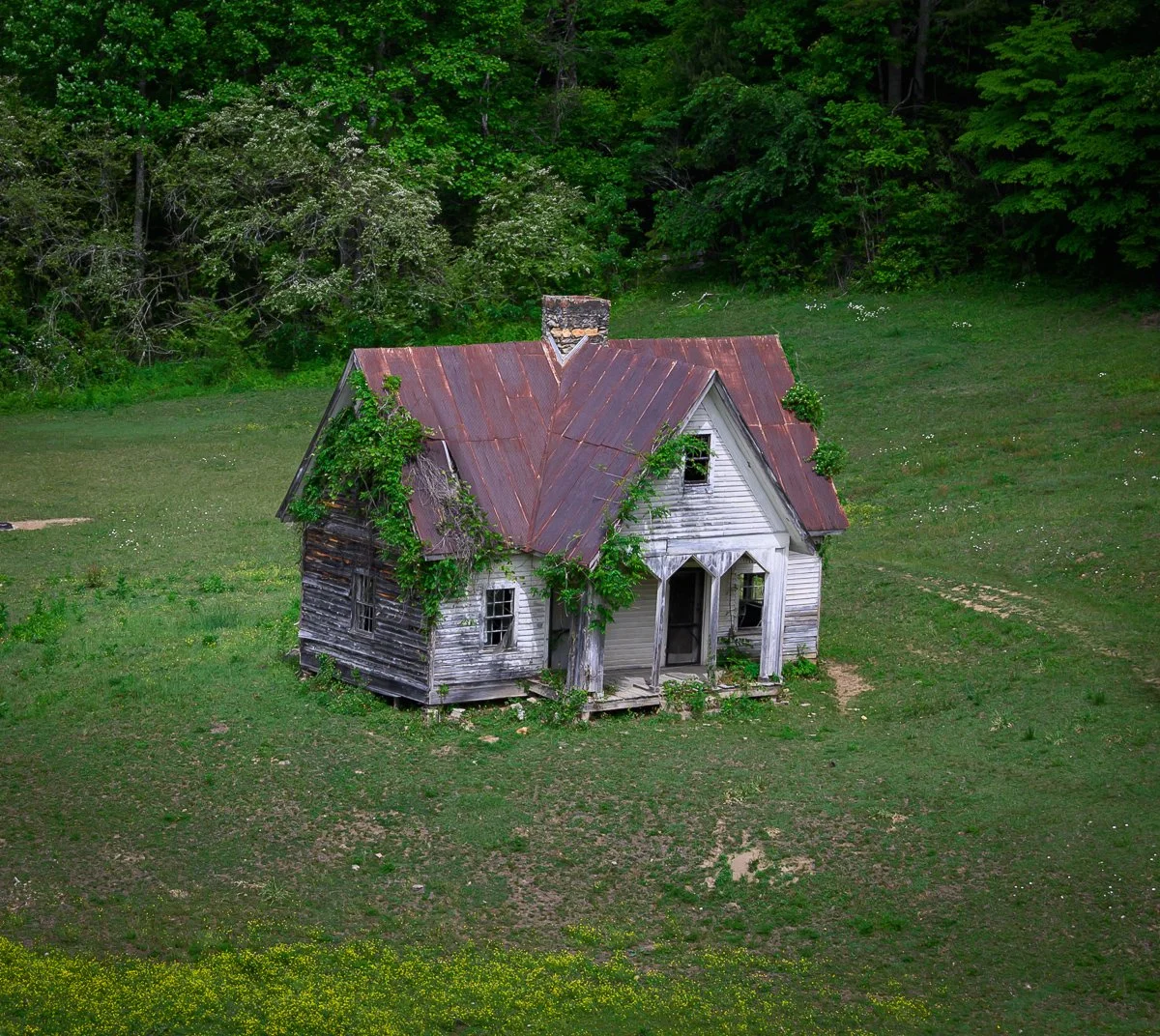 An old, abandoned house with a rusty metal roof and peeling white paint, surrounded by overgrown grass and trees.