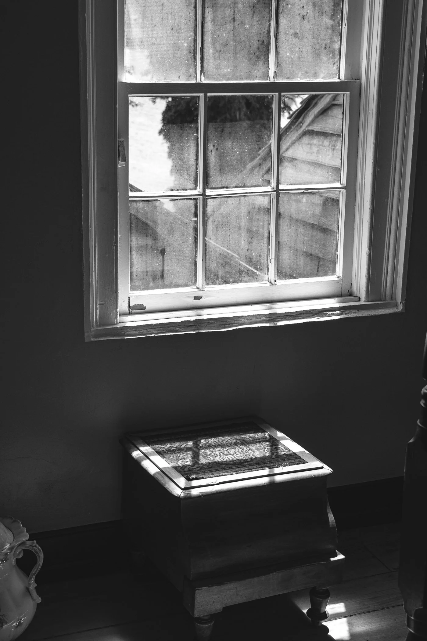Black and white photo of a window with sunlight casting shadows on a cushioned window seat below, and a part of an antique-style chair on the bottom left.