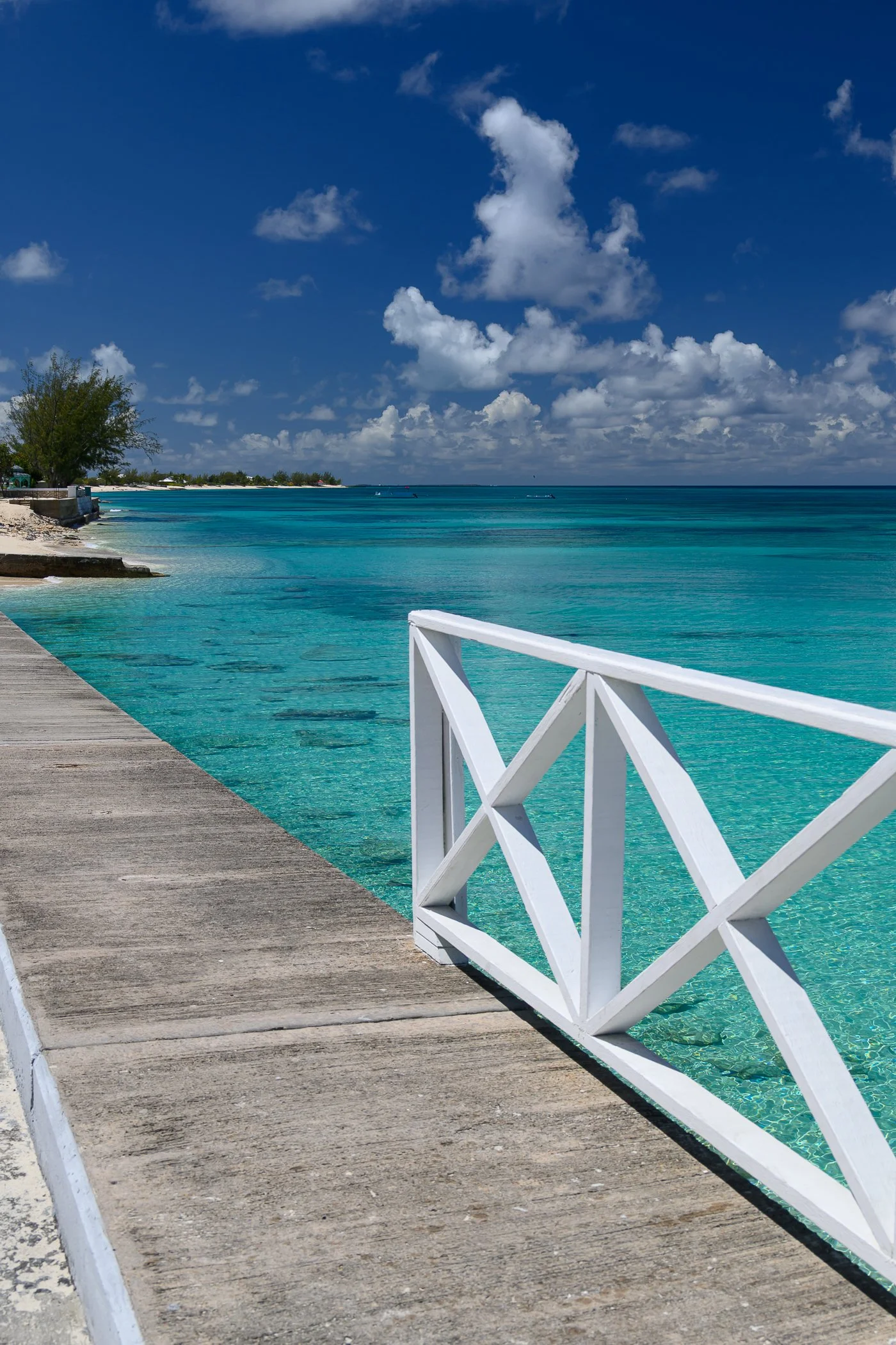 View of a beach with turquoise water, a concrete boardwalk, a white fence, green trees, and a blue sky with scattered clouds.