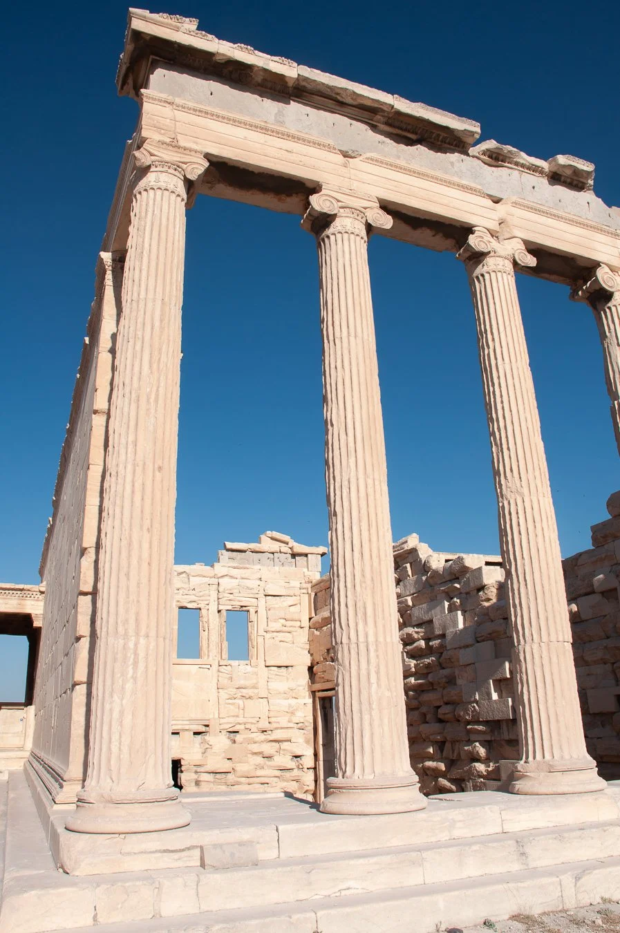 Ancient Greek ruins with tall, fluted columns and a partially collapsed structure against a clear blue sky.