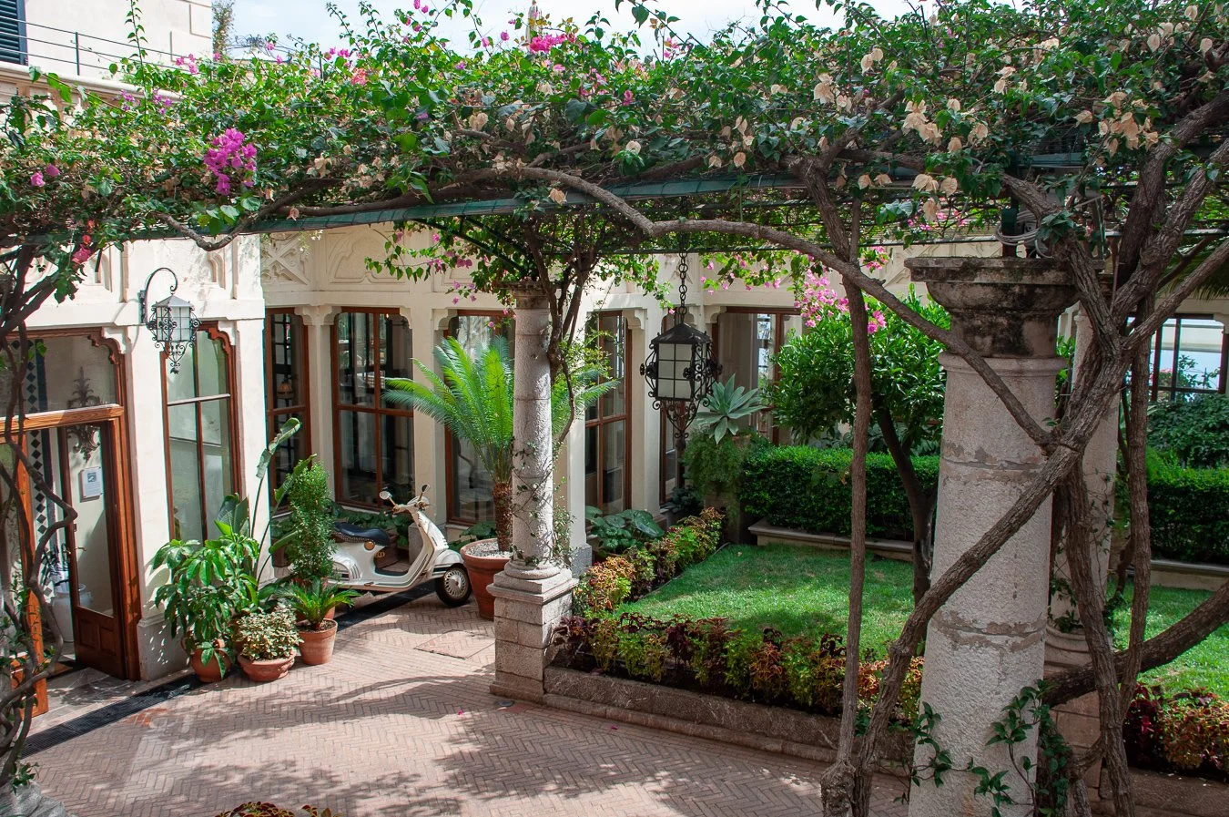 Courtyard garden with brick-paved flooring, potted plants, a small white scooter, and green plants, shaded by a large flowering vine on a pergola, surrounded by stone columns and ornate window frames.
