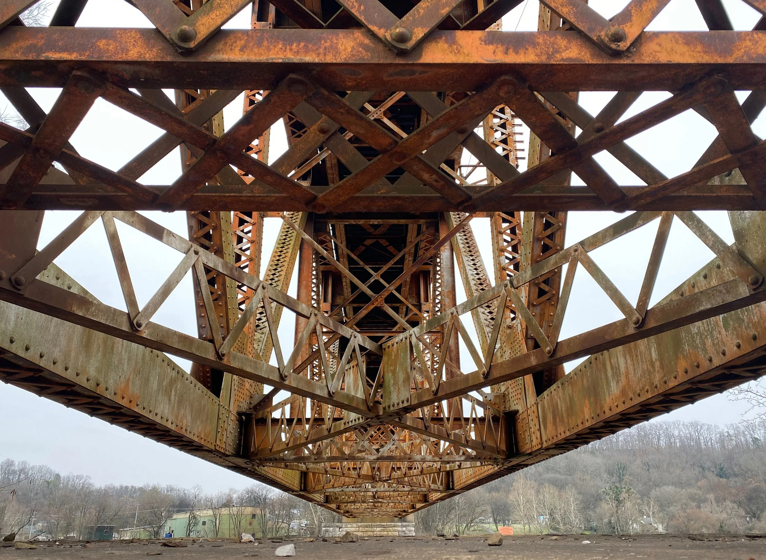 Underneath view of a tall, rusted metal bridge with a lattice structure, showing the underside and supporting beams. Trees and buildings are visible in the background.