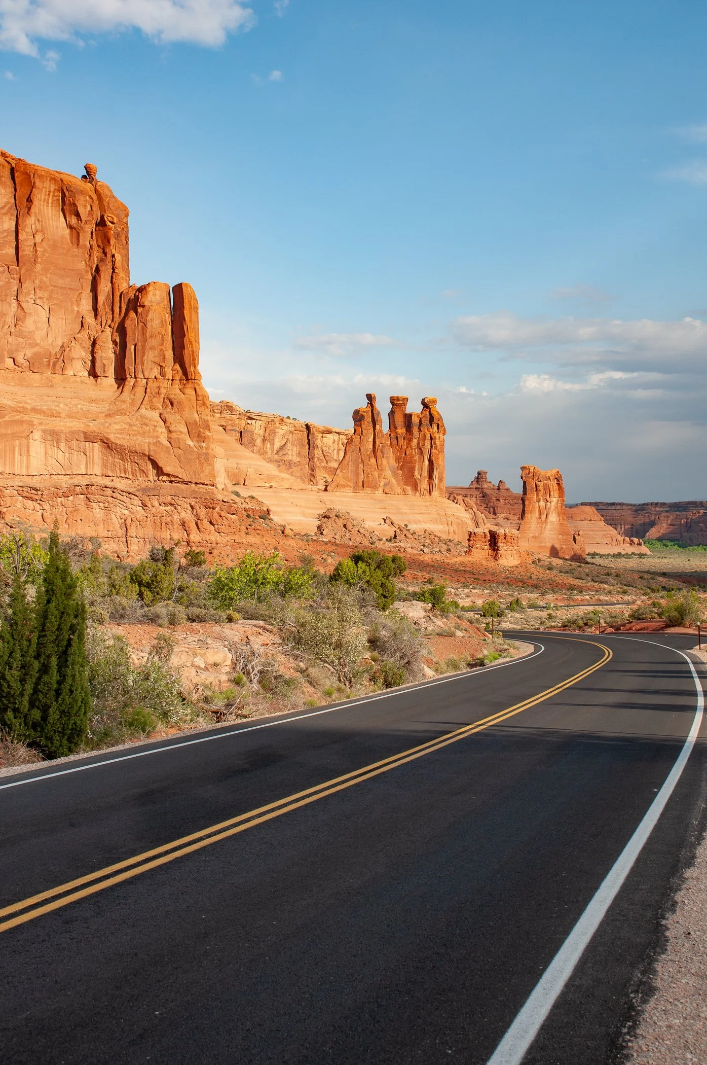 A winding two-lane road runs through a desert landscape with red rock formations and green shrubs, under a partly cloudy blue sky.