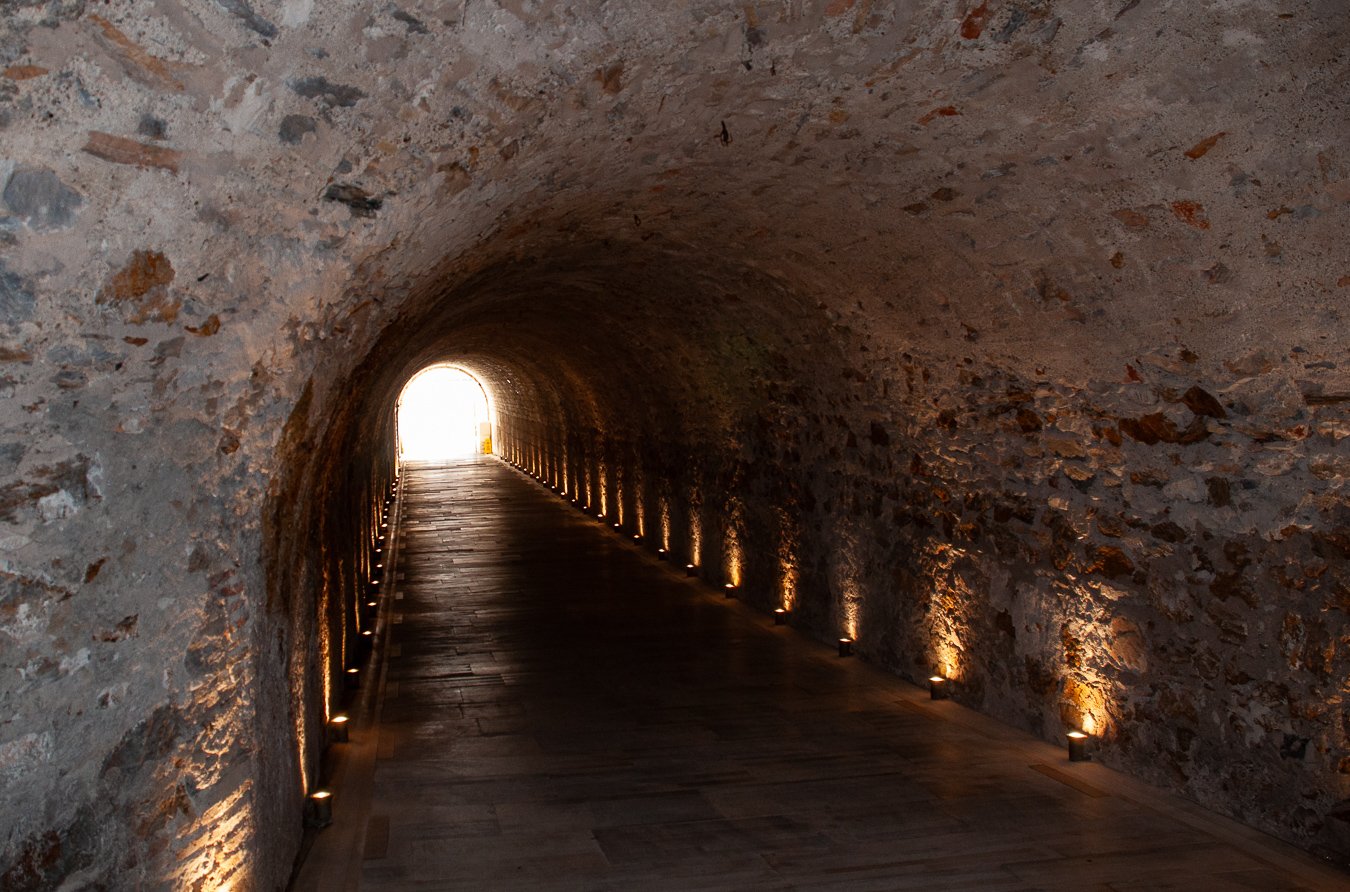 Underground tunnel with stone walls, lit by small lights along the sides, leading towards bright sunlight outside.