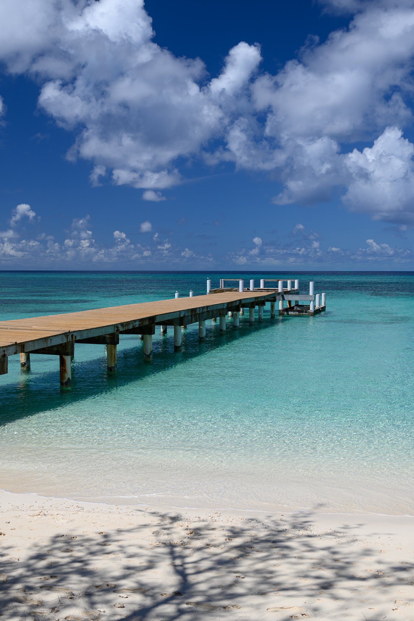 A sandy beach with clear turquoise water, a wooden pier extending into the ocean, blue sky with white clouds, and shadows of tree branches on the sand.