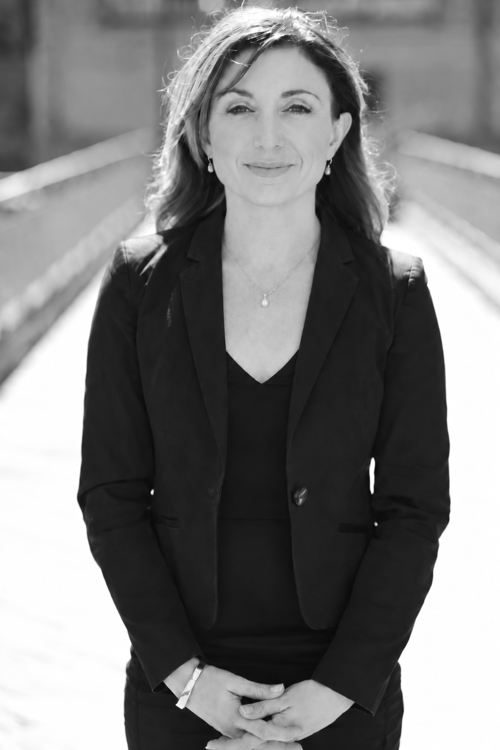 Black and white portrait of Corinne MacLellan, wearing a blazer and jewelry, standing outdoors on a sunlit bridge.