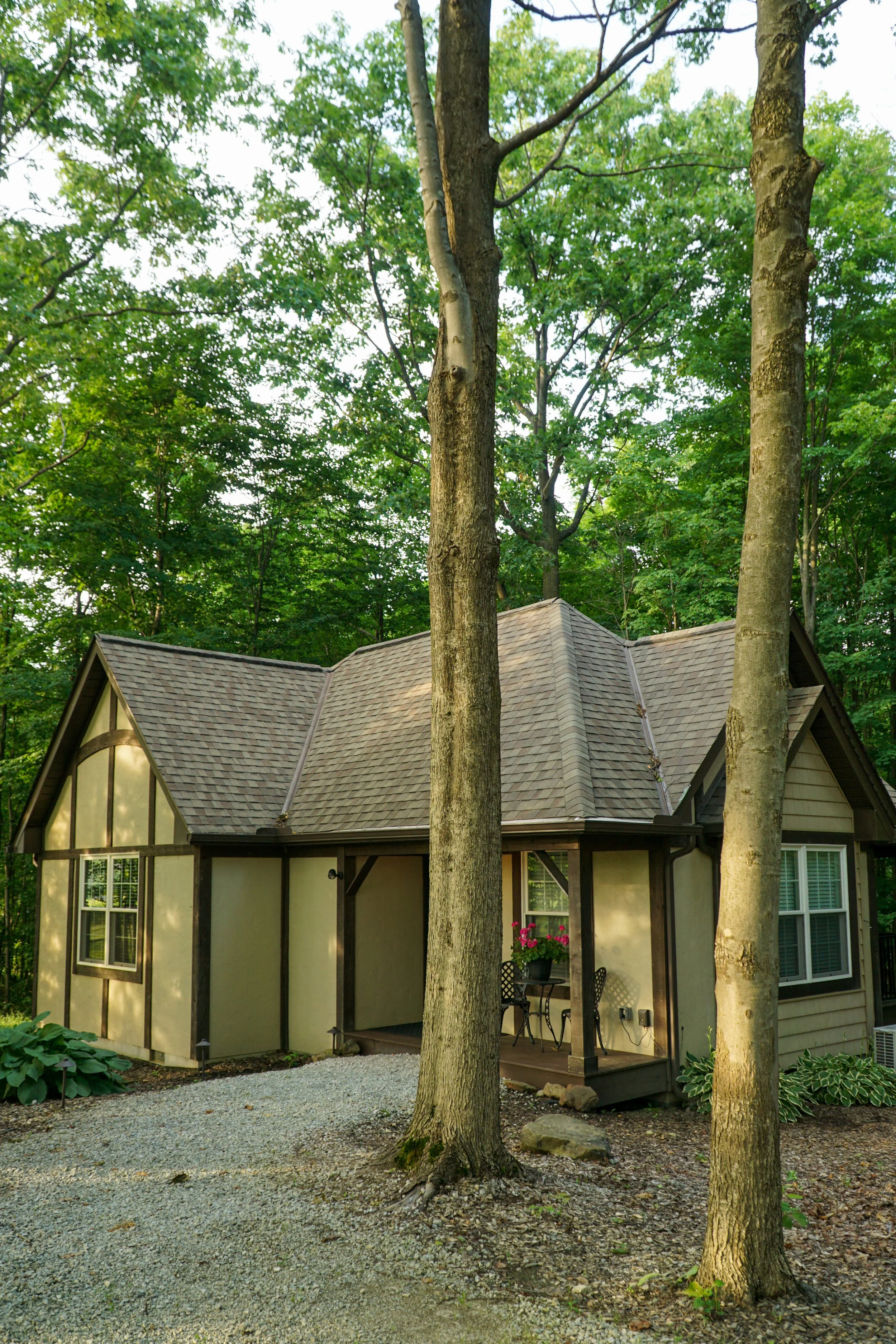 A house with a gabled roof, surrounded by trees, with a small porch with flowers, and a gravel pathway in the yard.