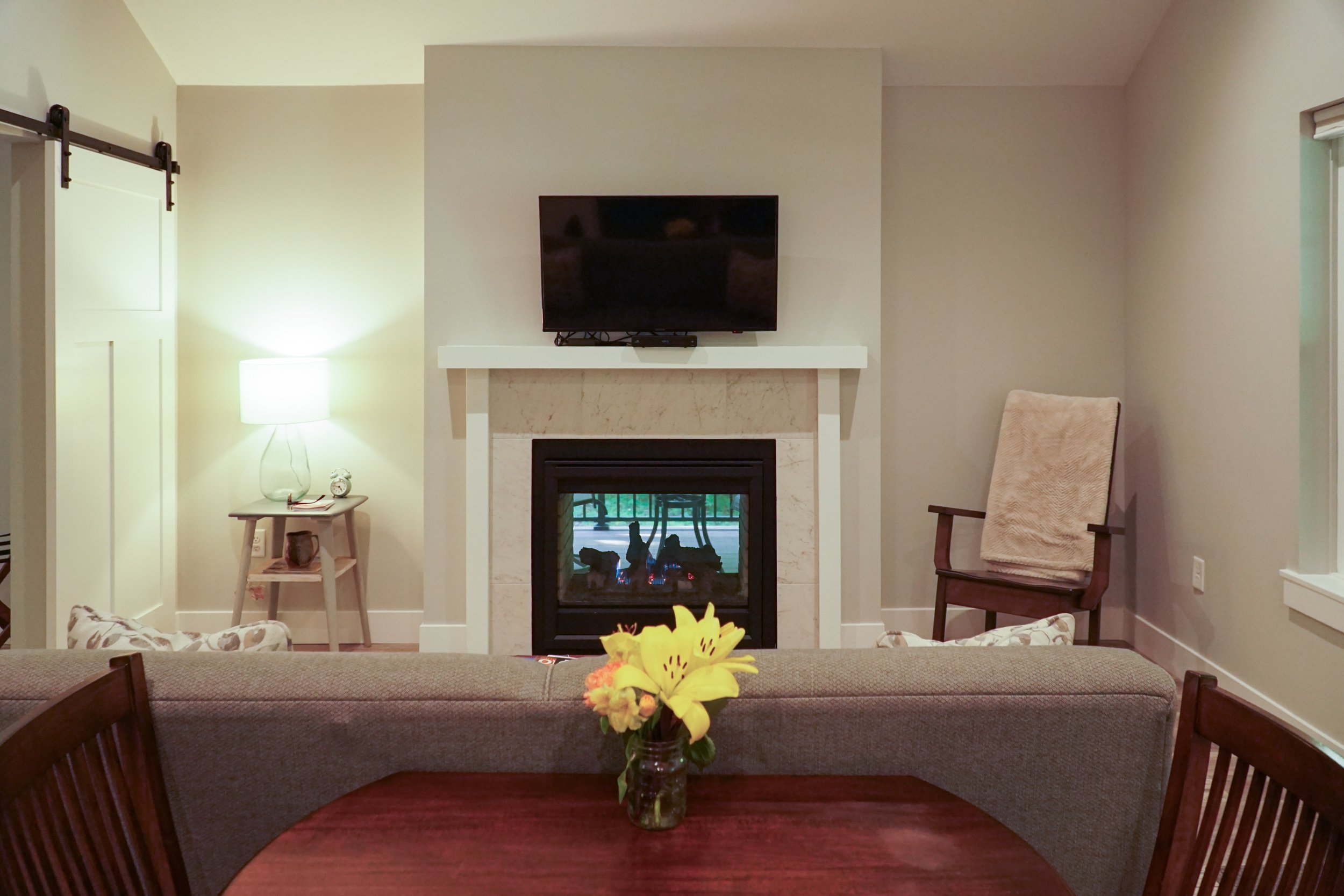 Living room with a fireplace, a mounted TV above it, a wooden dining table with a floral centerpiece, a beige armchair, a small side table with a lamp, and a sliding barn door on the left.