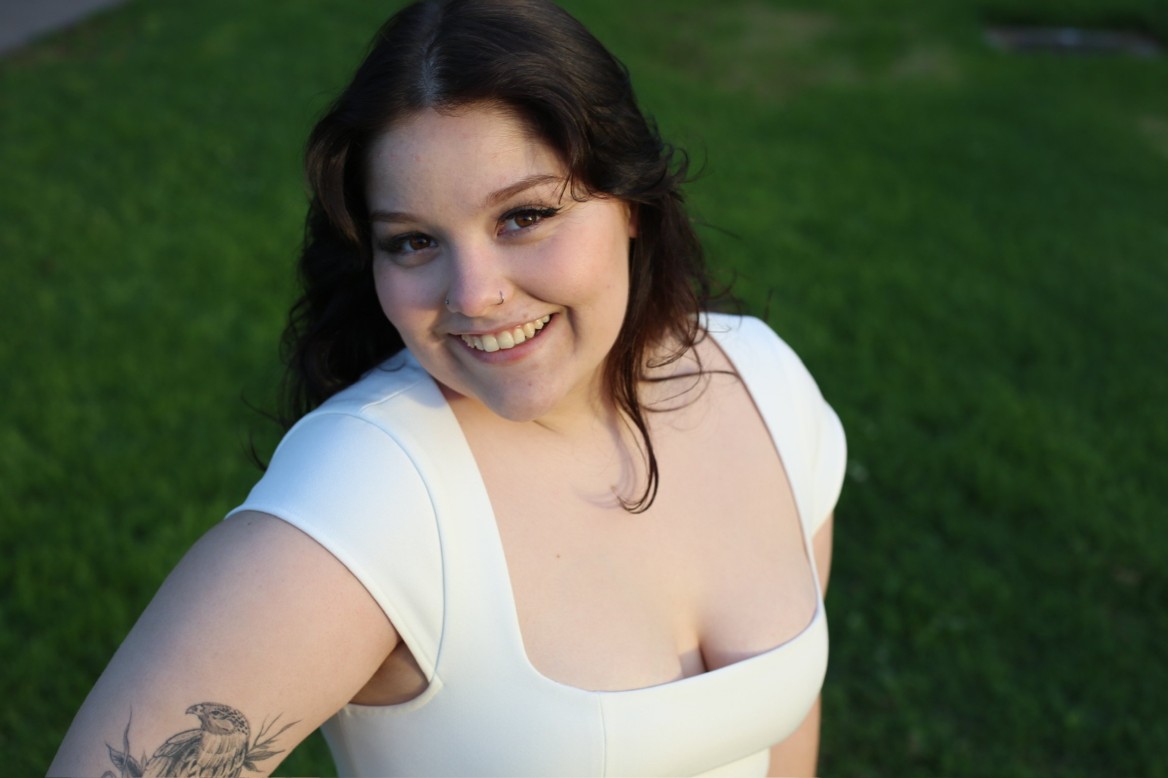 A woman with dark, wavy hair smiling outdoors, wearing a white top, with green grass background.