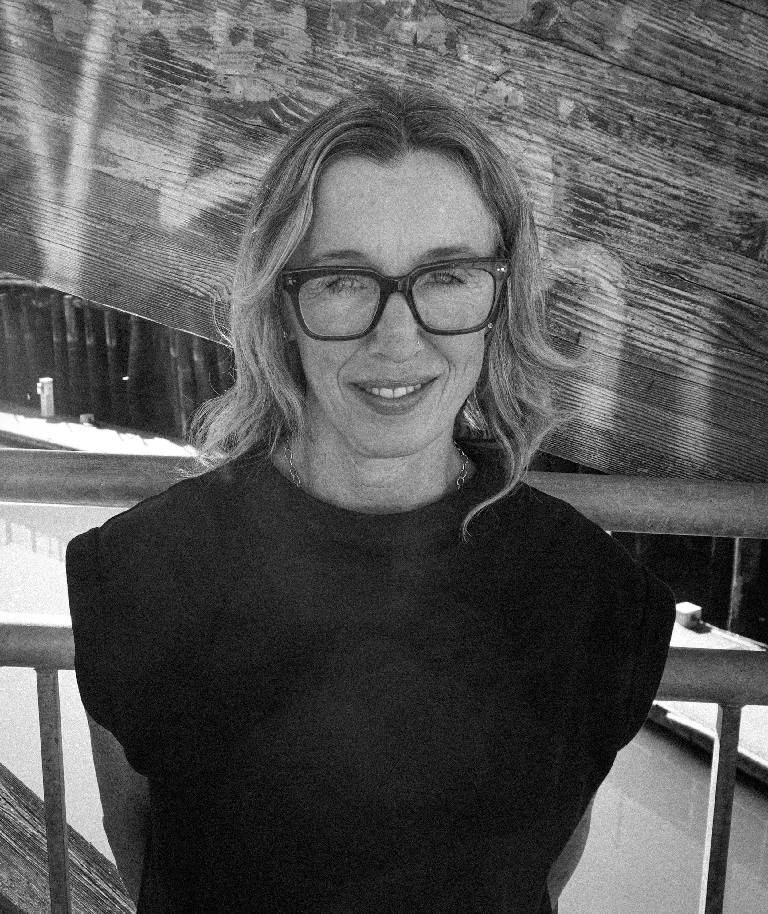 A woman with glasses and wavy hair taking a selfie outdoors with a wooden structure in the background.