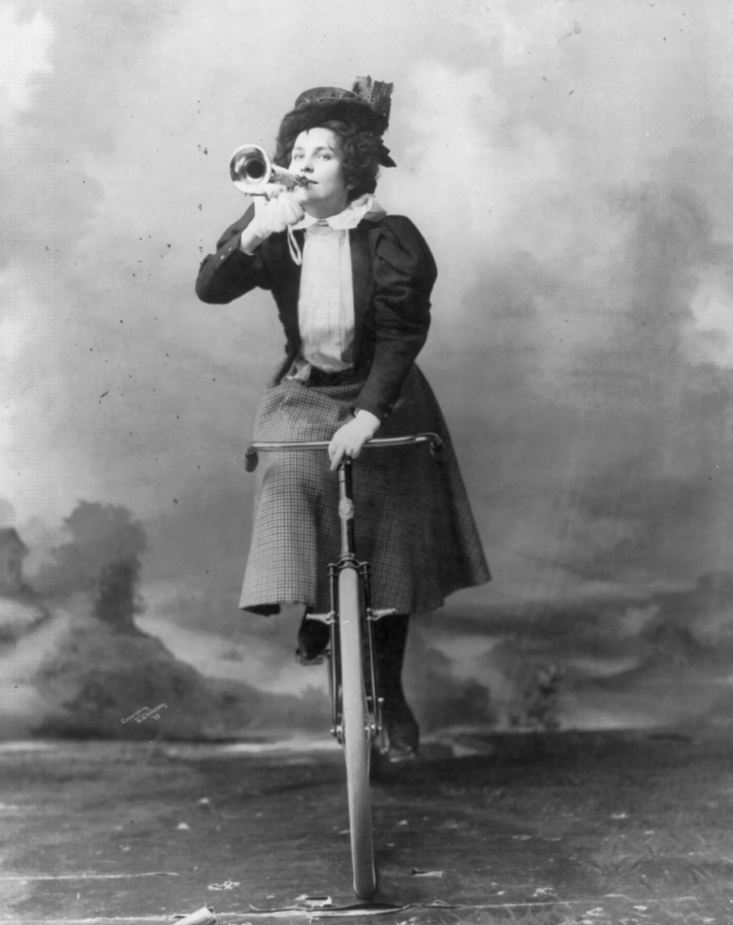 A black and white photograph of a woman riding a scooter. The woman is dressed in vintage-style clothing, wearing a hat with decorative elements, a dark jacket, a light blouse, and plaid pants. She is holding a small horn to her mouth as if to speak or shout. The background features a sky with some clouds, and the ground appears to be a flat, open space.