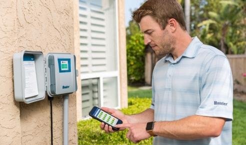 A man standing outside next to an irrigation clock reading data on his smartphone.