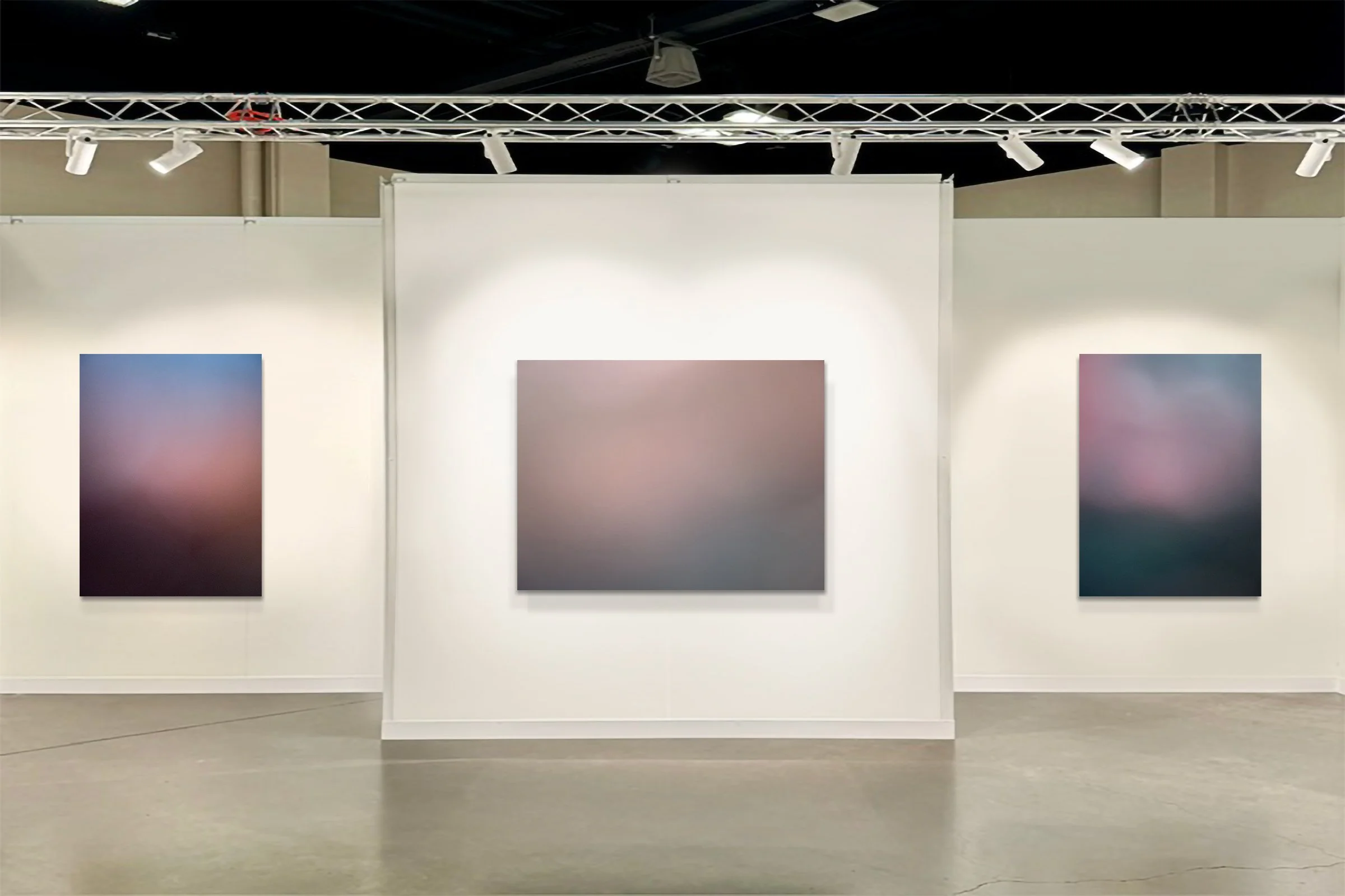 Three large scale abstract color field paintings hang on white gallery walls, styled as atmospheric landscapes featuring fields of color and luminous sunset skies.