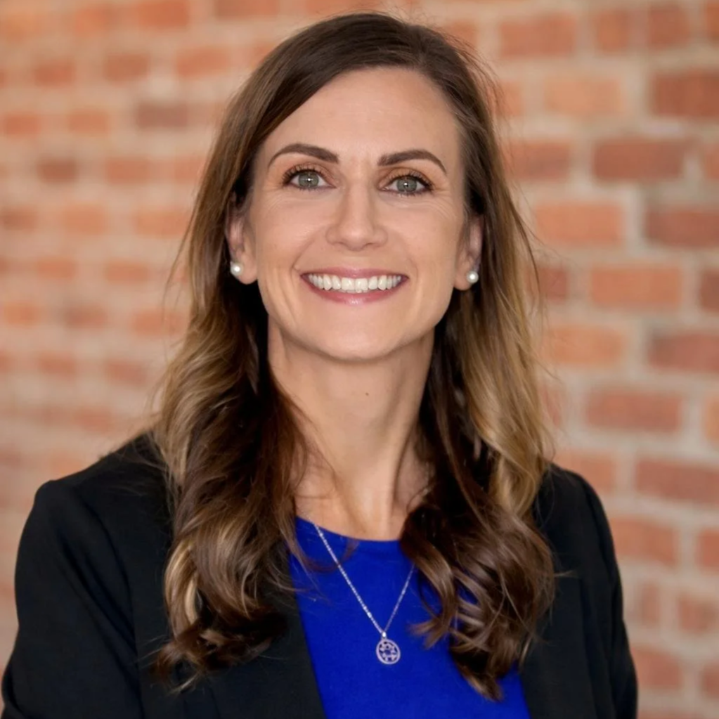 A smiling woman with wavy brown hair, wearing a blue top, black blazer, pearl earrings, and a silver pendant necklace, standing in front of a red brick wall.