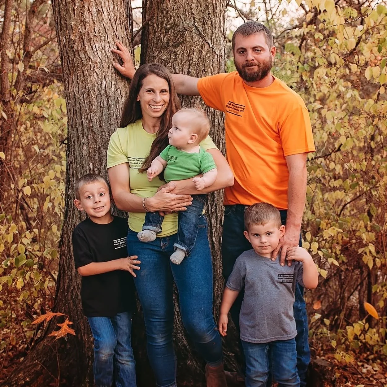 A family of six standing outdoors in front of a large tree with autumn leaves. The mother is holding a baby, and there are two young boys and a man, possibly the father, all posing and smiling.