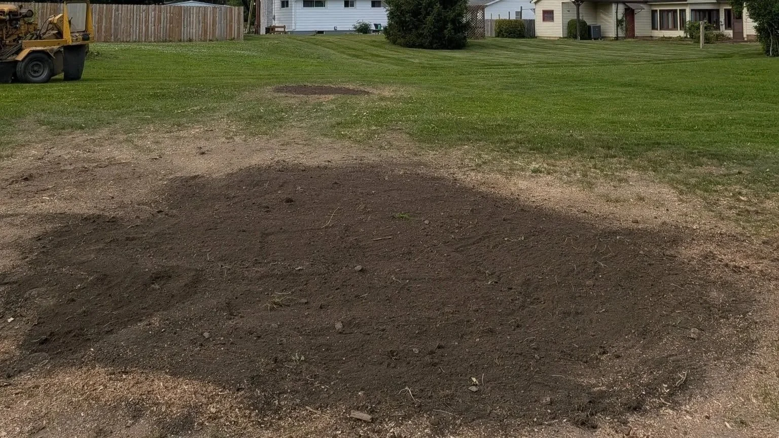 A backyard with patches of green grass and areas of bare soil. There is a wooden fence on the left, some houses in the background, and a construction vehicle on the left side.