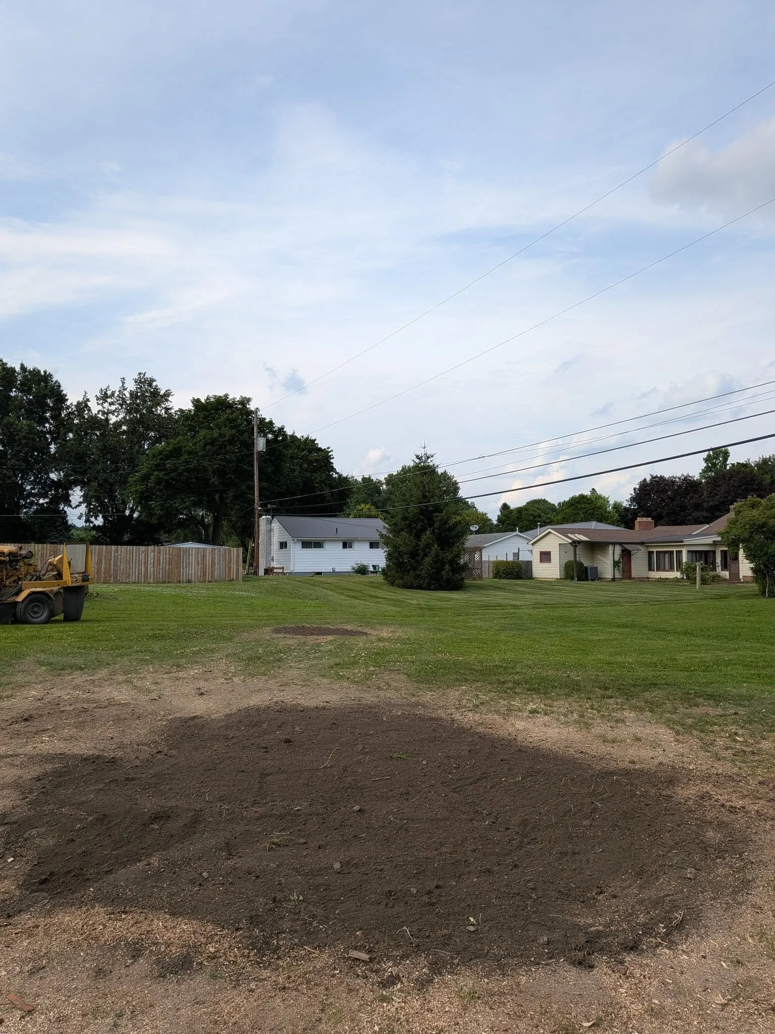 View of a backyard with a yard tractor, green grass, trees, houses, and power lines under a partly cloudy sky.