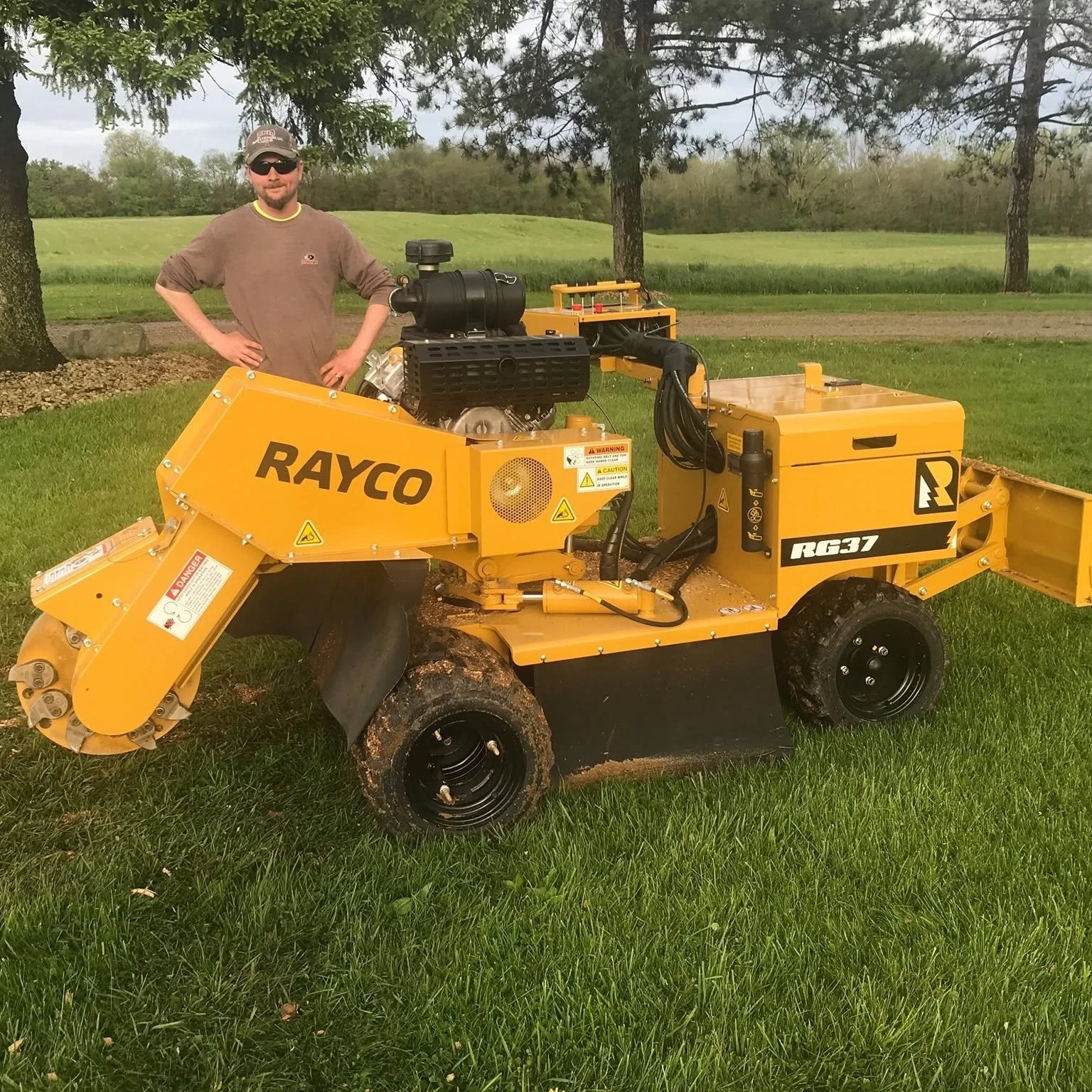 A man in sunglasses and a brown shirt standing next to a yellow Rayco RG37 stump grinder on a grassy area with trees and a field in the background.