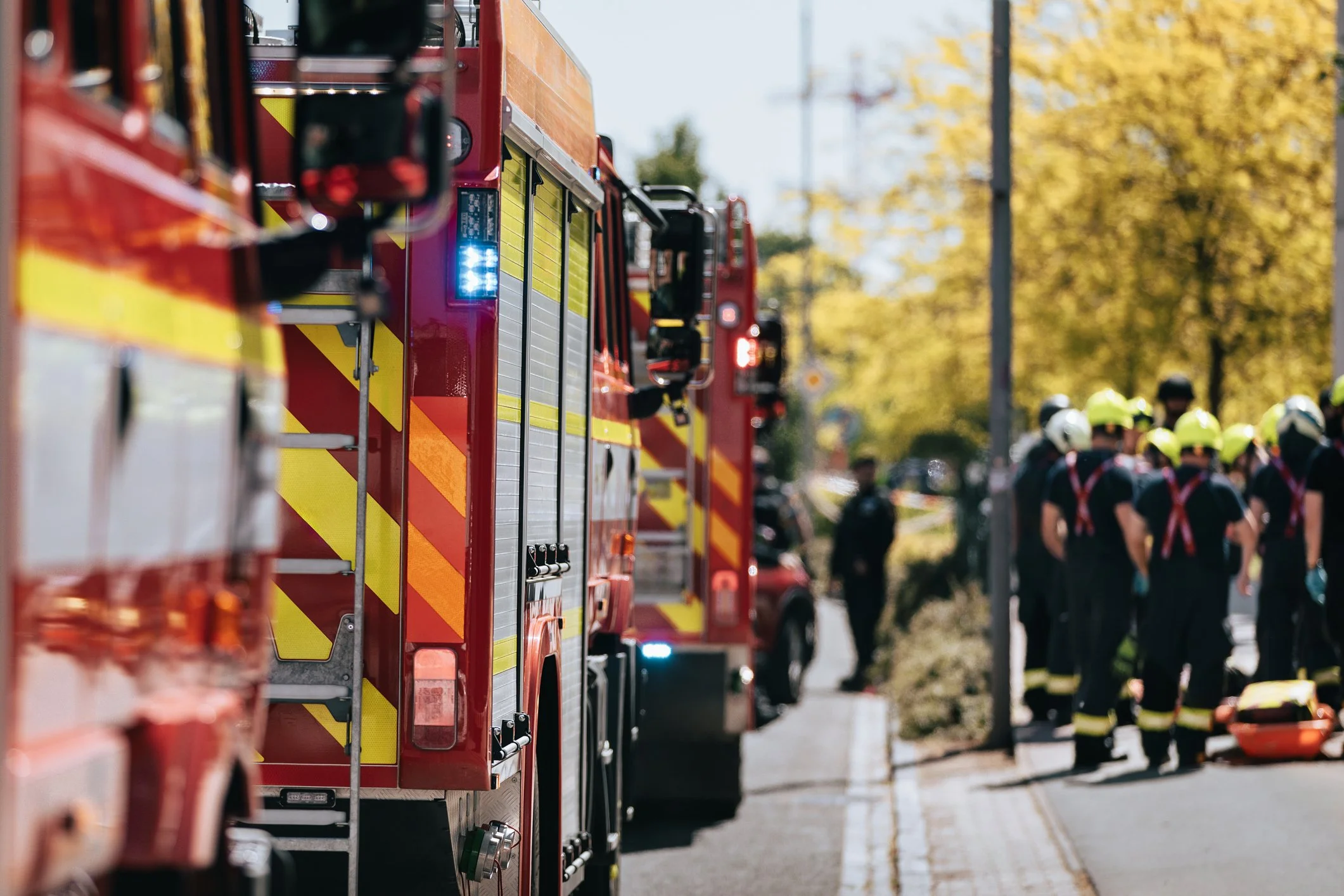 Fire trucks parked along the street with firefighters and emergency personnel in the background, some wearing helmets.