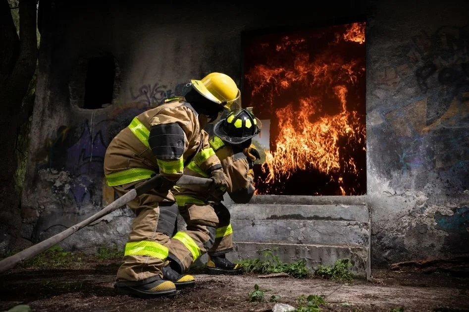 Two firefighters in full gear battling a fire inside a building, with flames visible through an open window.