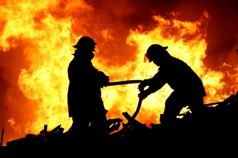 Silhouettes of two firefighters battling a large blaze with flames in the background.