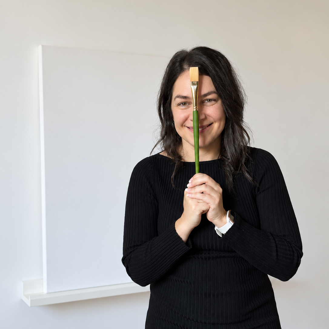 Femme souriante tenant un pinceau verticalement devant son visage, en direction du haut dans une pièce avec mur blanc.
