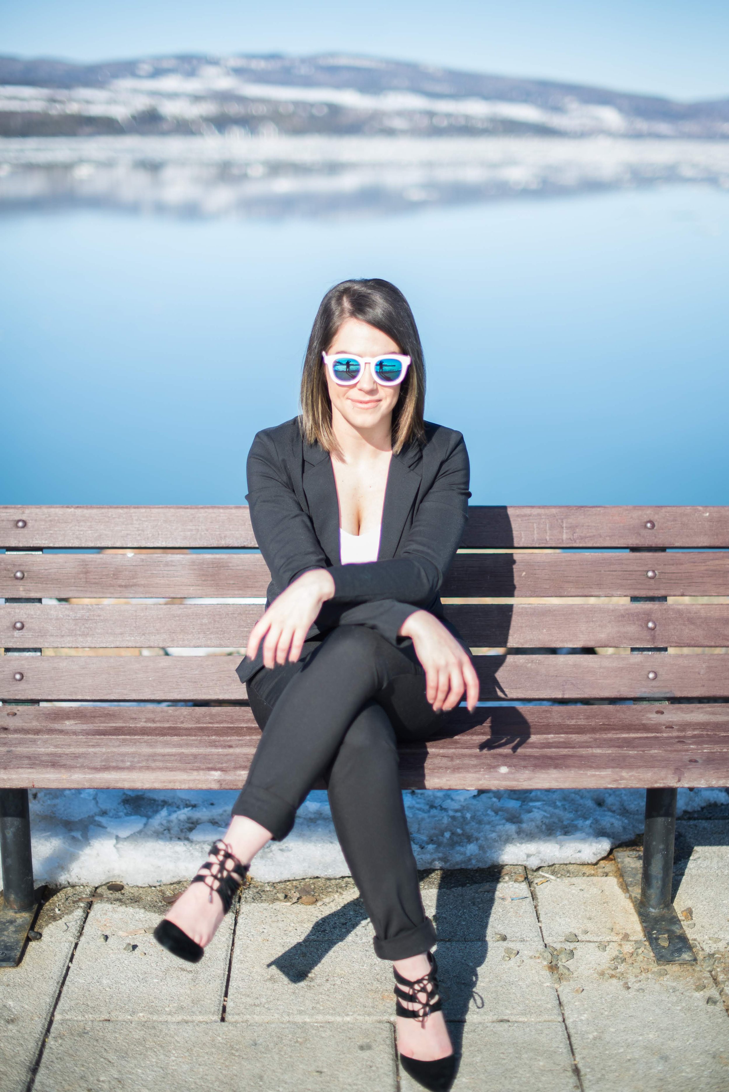 Une femme portant des lunettes de soleil, une veste noire et un pantalon noir, assise sur un banc en bois avec un paysage de lac et de montagnes enneigées en arrière-plan.