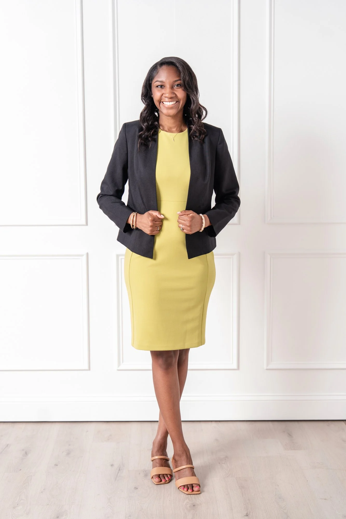 Georgia Registered Dietitian Nutritionist Destiney Mallett in a black blazer and yellow dresses standing indoors against a white paneled wall, smiling at the camera, with crossed legs and beige high-heeled sandals.