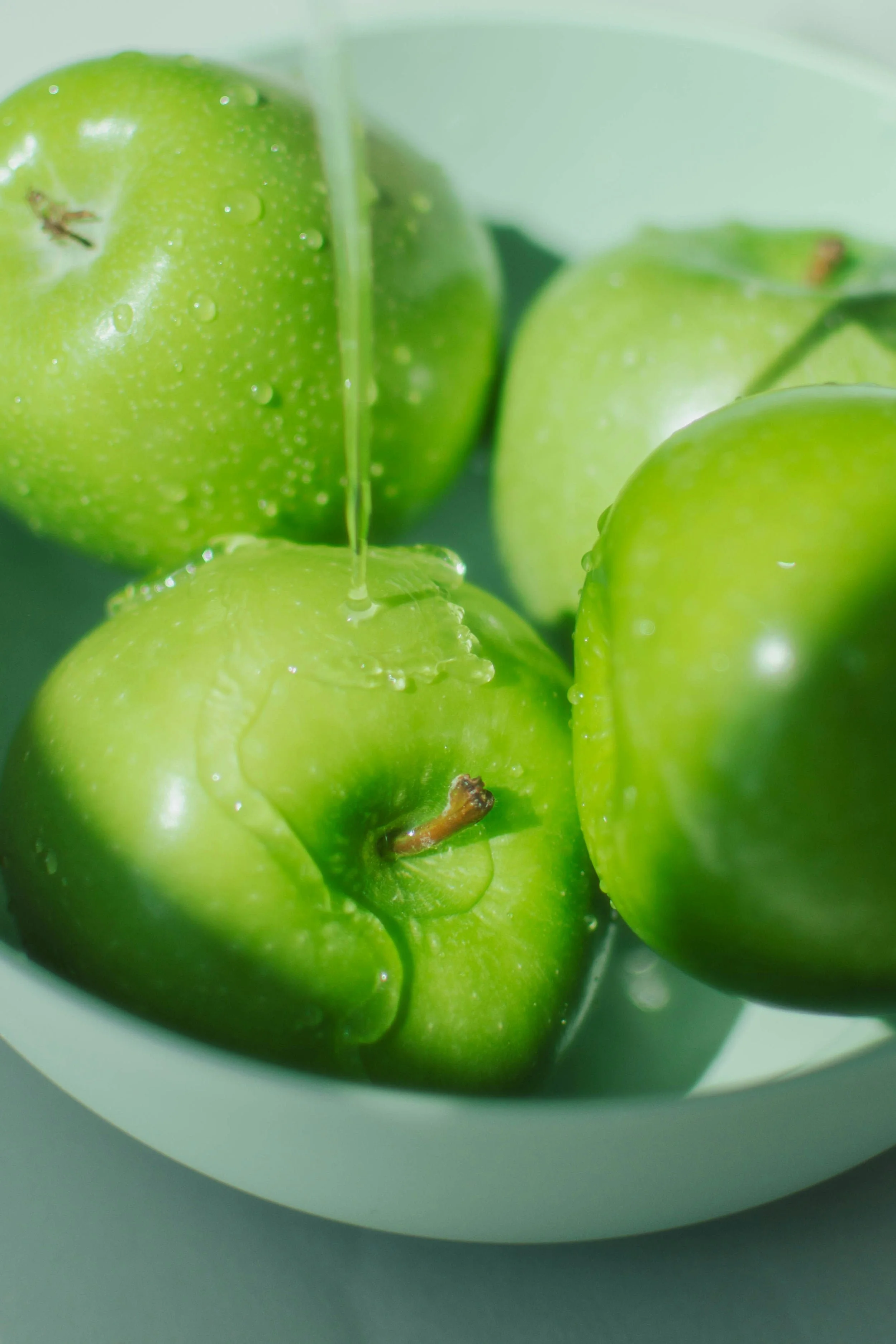 Close-up of three green apples with water droplets, one with a small blemish, inside a white container on a gray surface.